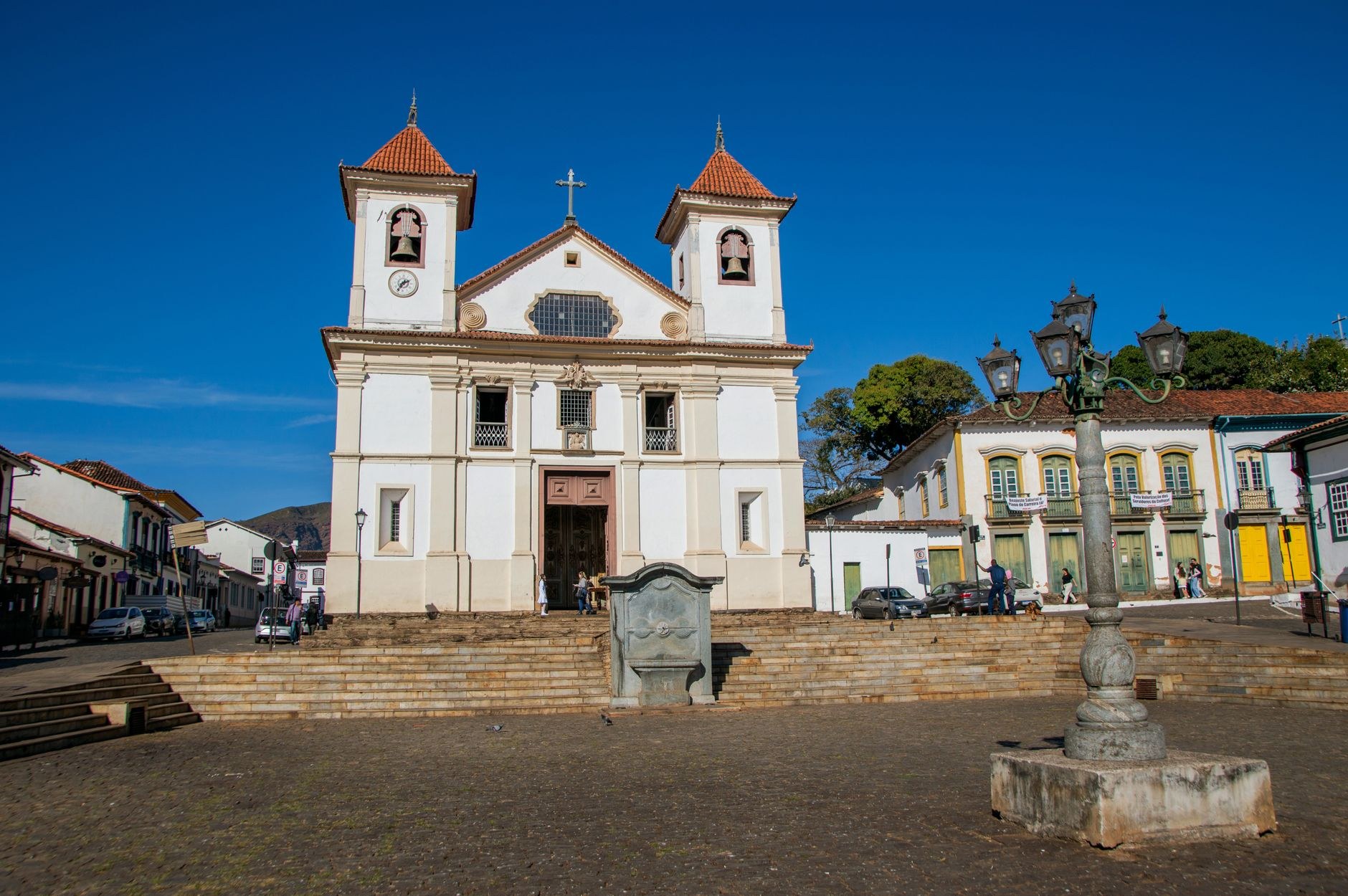 Igreja de arquitetura colonial em praça de pedra, cercada por casarios históricos, evoca o patrimônio das cidades históricas