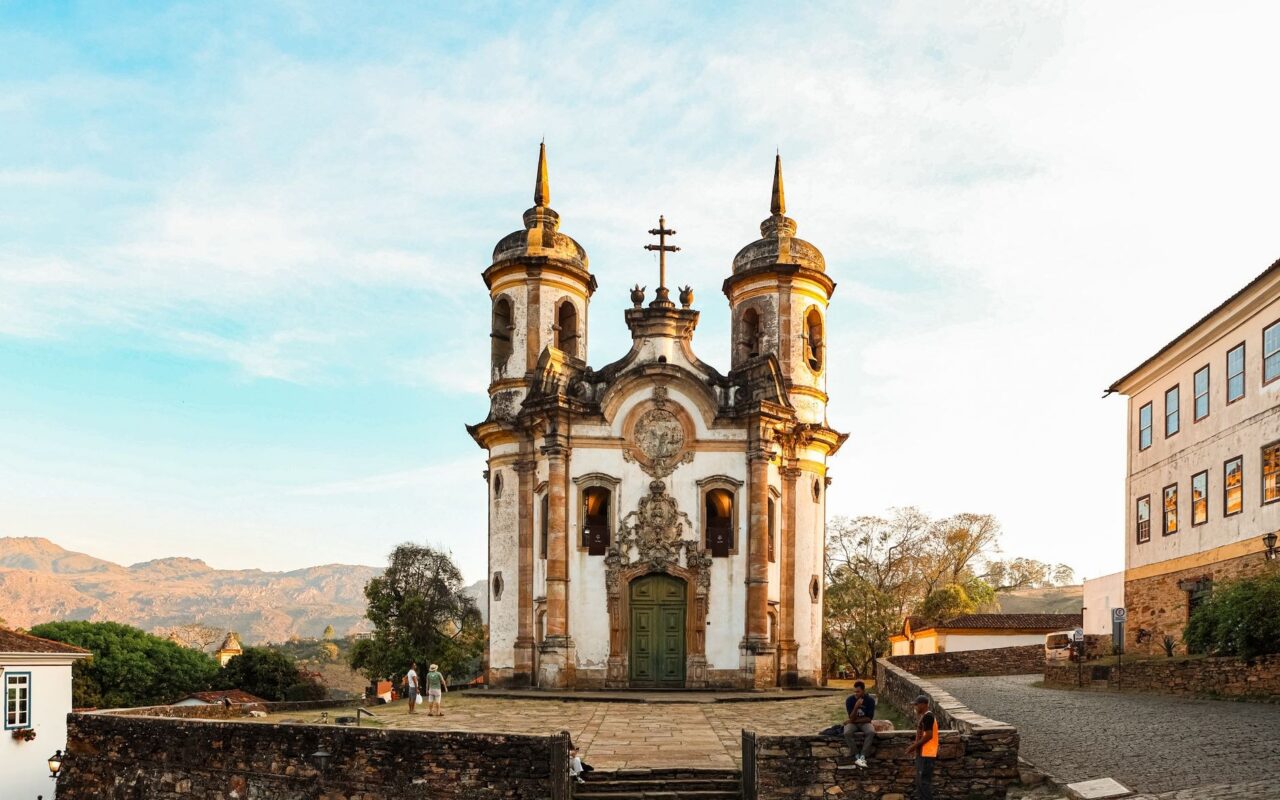 Igreja barroca ao entardecer, com ruas de pedra e casarios ao redor, evoca o patrimônio colonial brasileiro