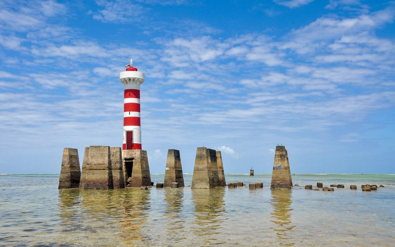 Pilares de concreto emergem da água rasa e sustentam um farol listrado, cartão-postal do litoral alagoano que atrai quem