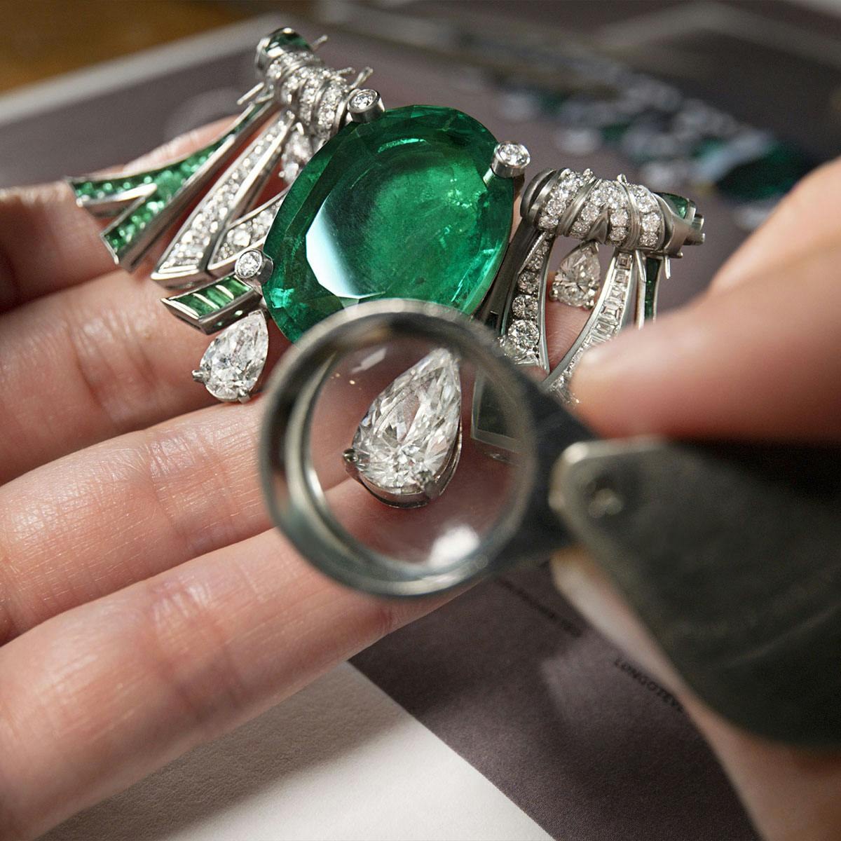 Artesão examina com lupa uma joia com esmeralda e diamantes, etapa de conferência do trabalho manual em joalheria de alto padrão