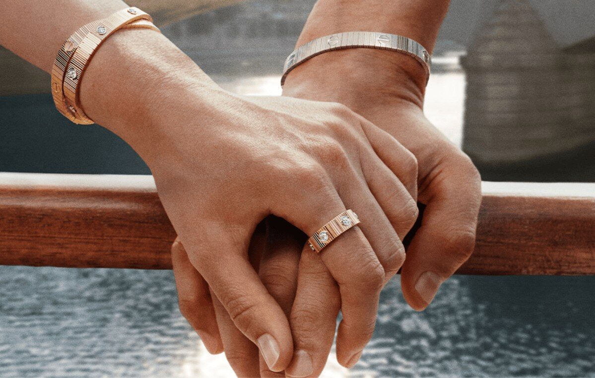 Mãos de mãos dadas exibem pulseiras e anel da linha LOVE Unlimited, da Cartier, com elos flexíveis e parafusos aparentes