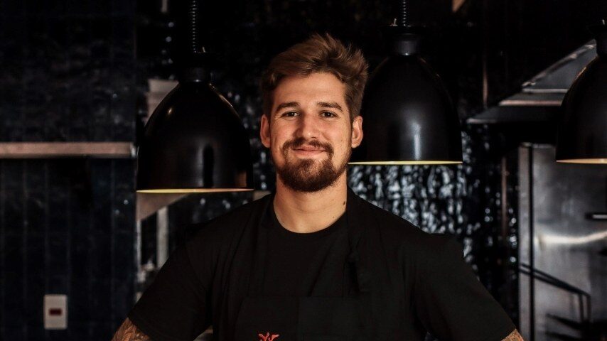 Chef de avental preto posa em cozinha profissional, em retrato de divulgação do Guia Michelin ligado à edição dos Jantar