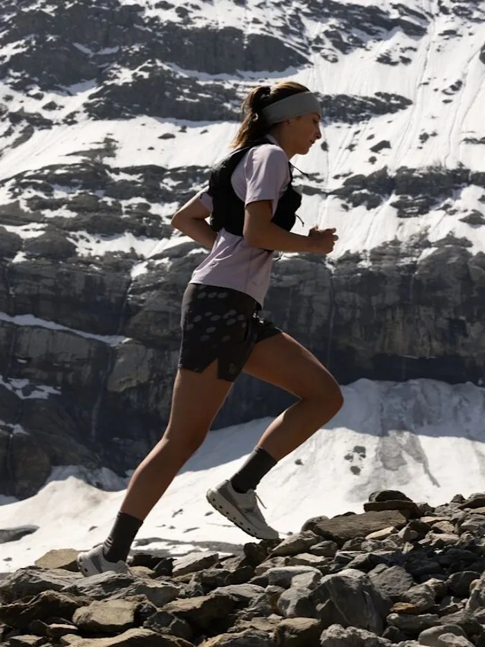 Corredora em trilha de montanha com neve ao fundo, calçando tênis de corrida On, categoria que originou a marca suíça