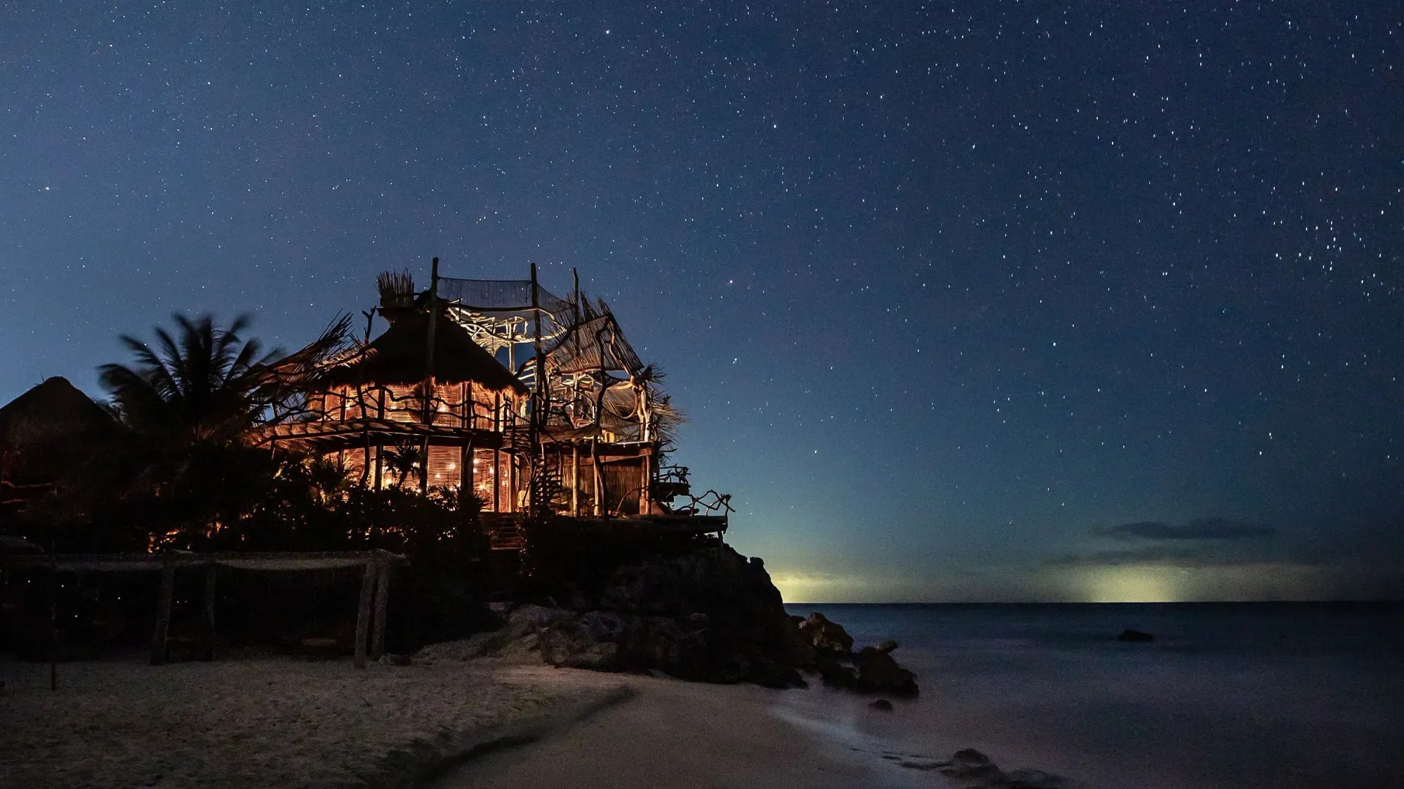 Ao anoitecer, a casa de madeira e palha acende entre palmeiras e rochas, com o céu estrelado sobre o Caribe